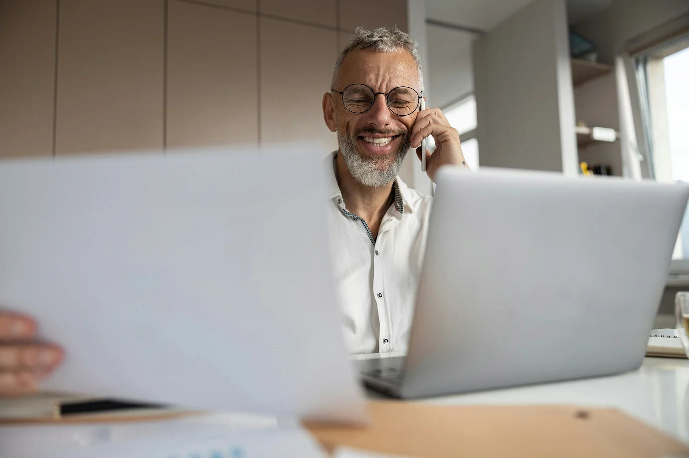 Lächelnder Geschäftsmann mit Brille telefoniert und prüft Unterlagen am Laptop im Büro